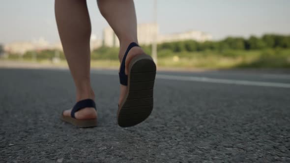 Female Feet in Black Sandals are Walking on an Asphalt Path