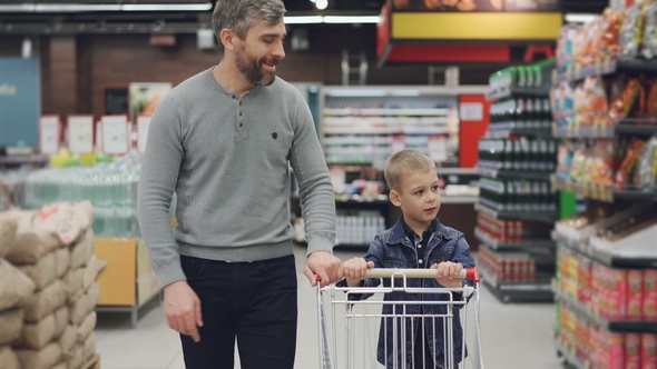 Dolly Shot of Young Family Father and Son Walking Through Food Store with Trolley Looking Around and
