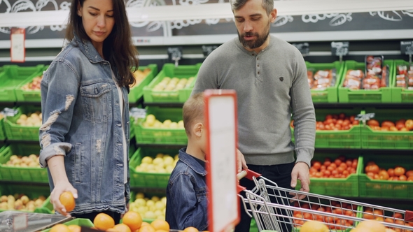 Young Family with Cheerful Child Is Walking Through Food Store with Shopping Cart and Choosing Fruit
