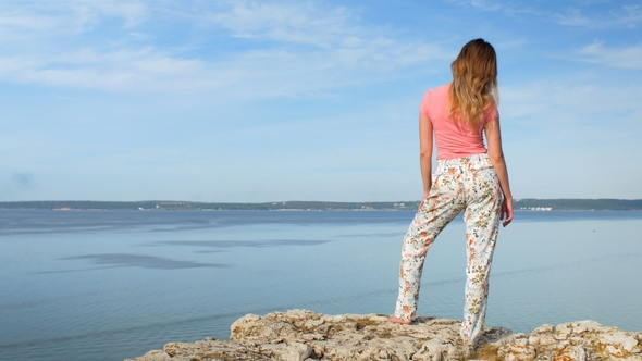 Young Slim Blonde with Wavy Hair Is Standing on Edge of Rock Over Wide Lagoon in Calm Sunny Weather