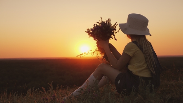 A Girl with a Bouquet of Wildflowers in Her Hands Is Looking at a Beautiful Sunset. Sits on a Hill