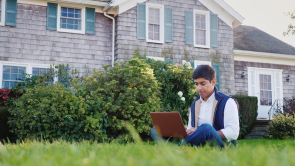 A Young Indian Man Works with a Laptop in His Backyard. Sits on a Green Lawn