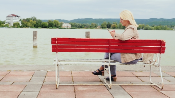 Woman Sits on a Bench on the Background of a Large Lake. Uses a Mobile Phone. Lake Balaton in