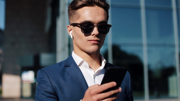 Young Businessman in Sunglasses Using Smartphone and Walking in the Street