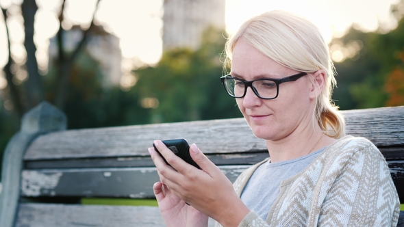 A Young Woman in Glasses Is Using a Smartphone. Sits on a Park Bench