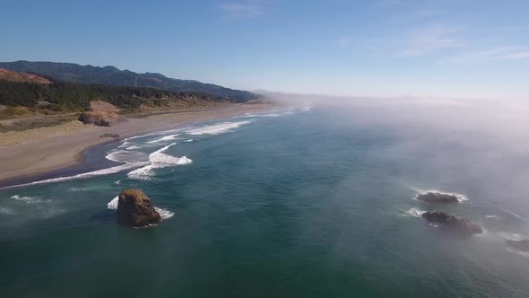 AERIAL: Flying over the Oregon coastline the waves crash against the shore as the fog rolls in.