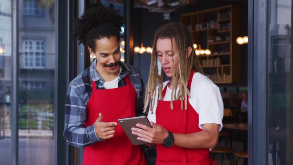 Two diverse male baristas wearing aprons standing in the doorway and using digital tablet