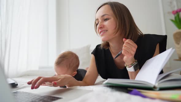 Young Mother Freelancer with Baby Son Sitting on Bed and Trying to Work at Laptop