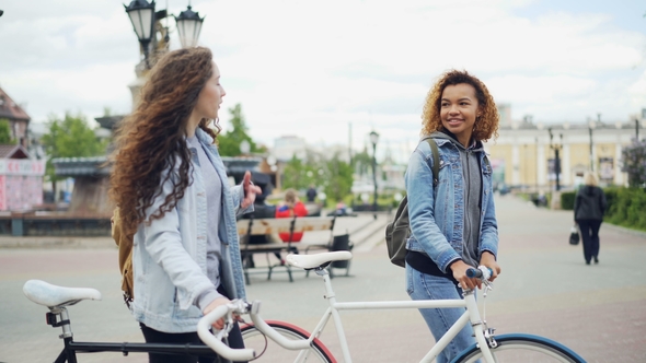 Cheerful Young Women Are Talking While Walking with Bikes Along Beautiful Street with Fountain and