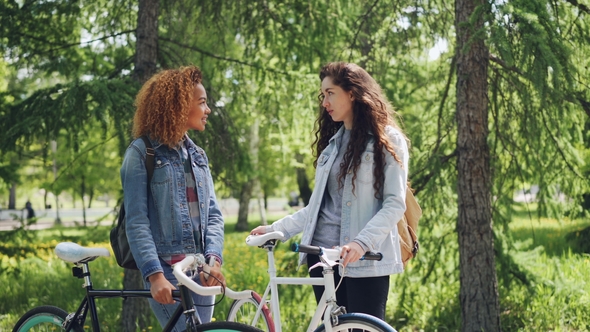 Two Female Bicyclers with Backpacks Are Talking Standing in the Park Holding Bikes