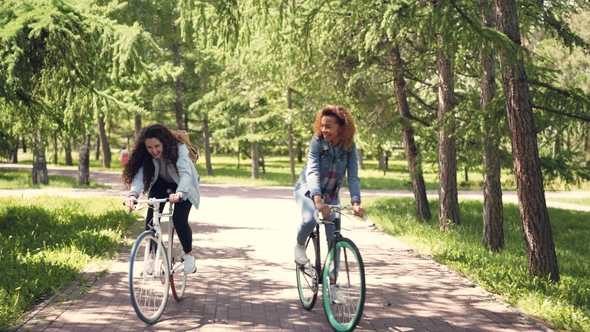 African American Girl Is Riding Bike in City Park with Her Caucasian Friend, Young Women Are Talking
