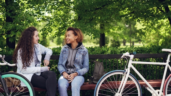 Cheerful Female Friends Are Talking Discussing News Sitting on Bench in Town Park with Bikes