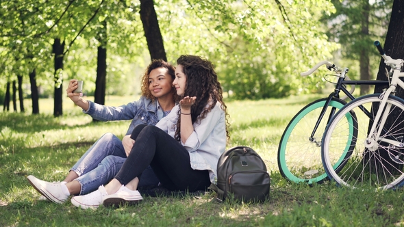 Young Woman and Her African American Friend Are Taking Selfie, Posing and Having Fun Sitting on Lawn