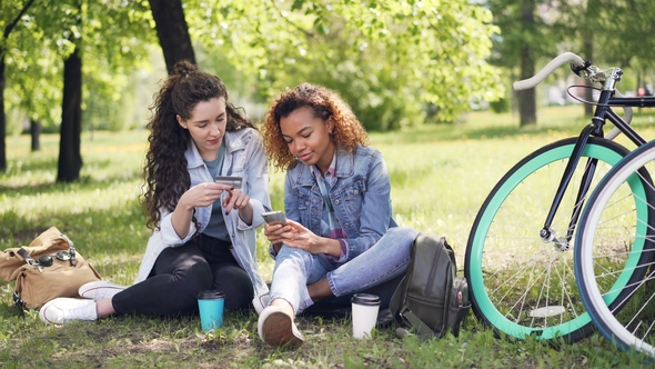 Two Young Women Are Paying with Card Using Smartphone Sitting in Park on Grass Holding Credit Card