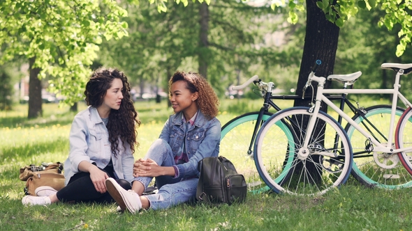 Cheerful Young Women Caucasian and African American Are Chatting in Park While Resting After Riding