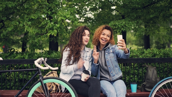 Female Friends Are Taking Selfie with Smartphone Sitting on Bench in Park Holding Coffee, Young