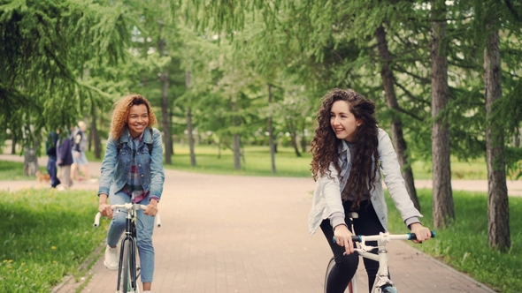 Dolly Shot of Happy Female Students Friends Riding Bikes in Park, Looking Around and Laughing