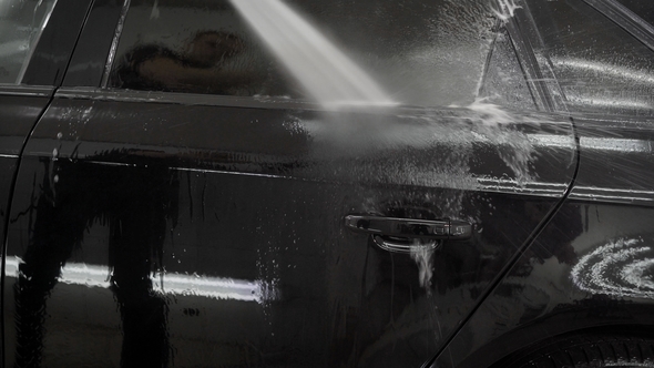 Man Is Pouring By Water From a Hose Black Automobile in a Garage, Washing Off Soapy Foam From a Body