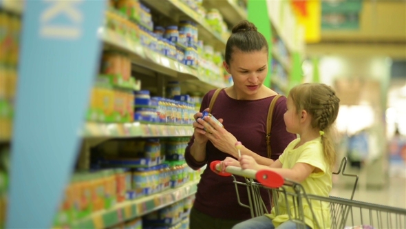 Cutie Girl with Caucasian Appearance Is Eating Lollipop and Sitting in Supermarket Trolley. Mother