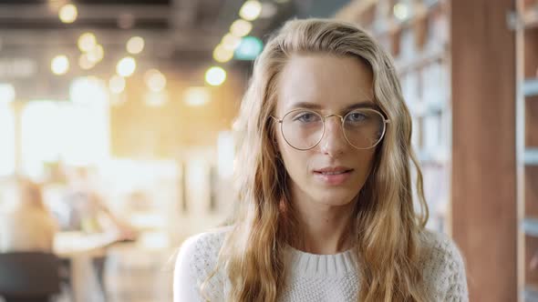 Portrait of Serious Clever Girl in Eyewear Looking at a Camera in a University Library Background. A