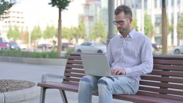 Busy Young Adult Man Using Laptop Sitting Outdoor on Bench