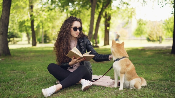 Cheerful Female Student Is Reading Book in the Park Sitting on Lawn and Caressing Beautiful Dog, Pet