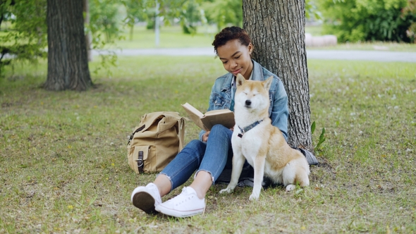 Cheerful Mixed Race Girl Dog Owner Is Reading Book and Stroking Her Cute Puppy Sitting on Grass in