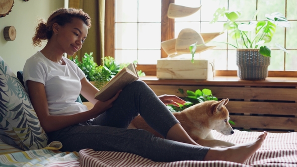 Attractive Girl Is Reading Book and Stroking Beautiful Dog Lying on Bed