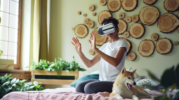 African American Girl Is Using Virtual Reality Glasses Sitting on Bed at Home, She Is Moving Hands