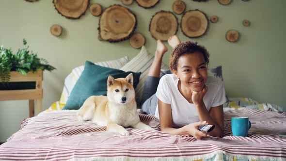 Cheerful Mixed Race Student Is Watching TV Holding Remote and Pressing Buttons Choosing Television