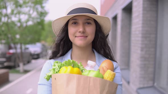 A Young Attractive Woman in a Denim Jacket and Hat Carries a Grocery Bag While Having a Good Mood