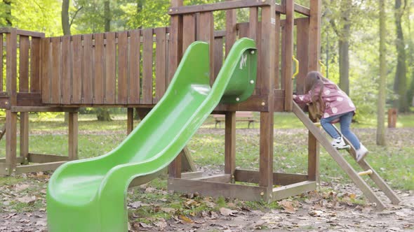 Cute Little Caucasian Girl Slides Down a Slide and Runs Away at a Playground in a Park