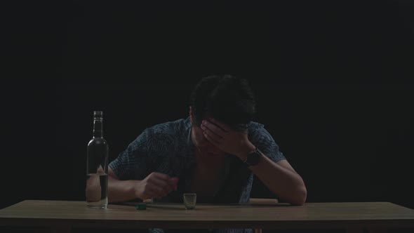 Drunk Asian Man Crying And Pouring Vodka In A Shot Glass Before Drinking In Black Background