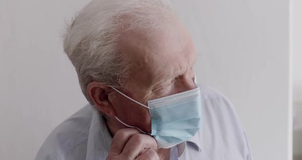 Senior Man in Mask Looking Out Window and at Camera During Lonely Selfisolation