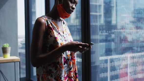 African american woman with face mask around her neck using smartphone at modern office