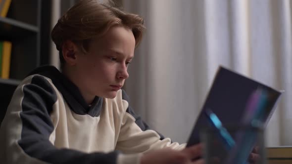 Closeup of Focused Serious Pupil Boy Reading Paper Book Sitting Alone at Desk in Light Children Room