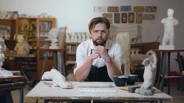 Sculptor with Beard in Glasses Sitting at His Workplace in the Studio