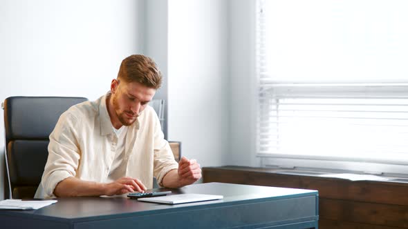 Thoughtful guy manager with beard sums up administrative expenses on calculator