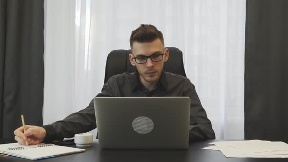 Male financier working on laptop in office takes note in notebook in front of laptop