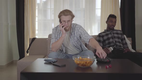 Handsome Bearded Man Sitting on the Sofa Talking By Cellphone While His African American Friend