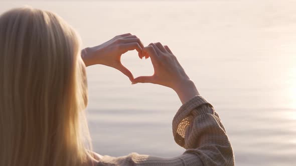 A Romantic Woman Stands By the Water and Shows a Heart with Her Hands at Sunset