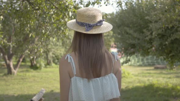 Back View of Cute Young Woman in Straw Hat and Long White Dress Walking Through the Green Summer
