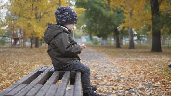 Lonely Small Boy Sits on the Bench with Phone in Hands