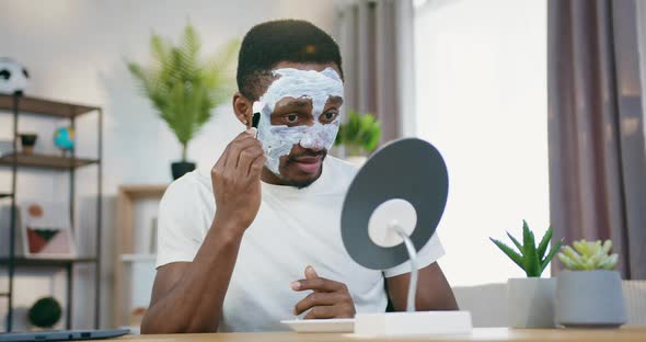 Black-Skinned Guy Looking Into Small mirror when Putting on Moisturiser Mask on face