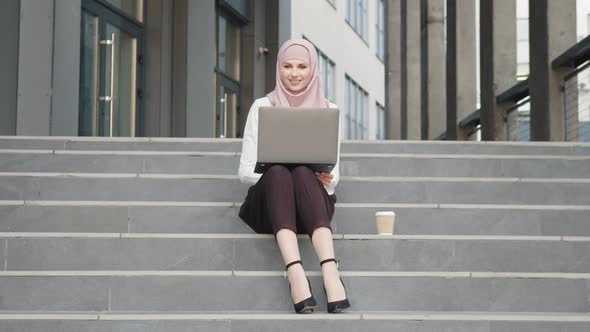 Smiling Young Woman in Hijab Woking on Wireless Laptop While Sitting on Stairs