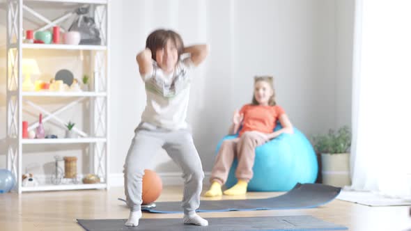 Cheerful Caucasian Brother Doing Squats As Blurred Sister Counting Sitting on Bag Chair