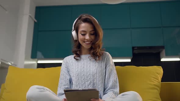 Cheerful Young Lady with Earphones Listening Nice Music, Sitting on the Cozy Kitchen Sofa