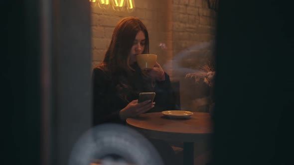 A Beautiful Young Girl Drinks Coffee in a Cafe and Uses a Mobile Phone