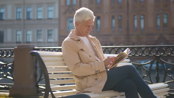 Elderly Casual Man Having Rest and Reading Book on Bench in City Center