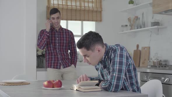 Portrait of Concentrated Caucasian Man Sitting at the Table and Reading As His Twin Brother Talking